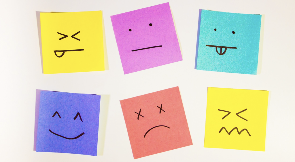 six sticky notes, all different colors are stuck on a flat, plain surface. In black marker, each note has a stick figure face reflecting different emotions: happy, angry, confused, etc.
